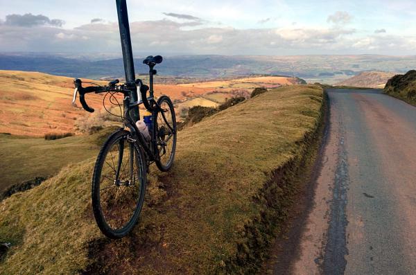 A touring bicycle parked beside a narrow country road on a hillside with expansive views across rolling Welsh valleys and distant mountains under a cloudy sky during golden hour light.