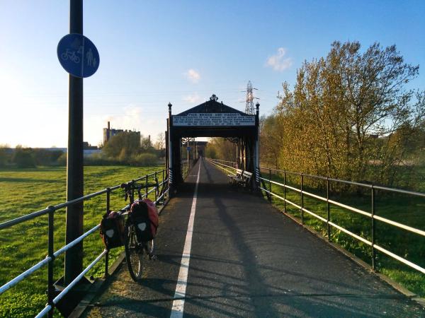 A touring bicycle with red panniers parked on a cycle path approaching a Victorian railway viaduct with an ornate entrance plaque under bright blue sky. A blue cycle route sign is visible on the left, and the path leads through the viaduct tunnel with green fields and trees on either side.