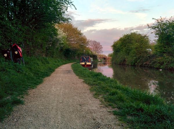 A peaceful canal towpath scene at sunset with a red and black narrowboat moored alongside the waterway, surrounded by lush green vegetation and trees with spring foliage. The gravel towpath stretches into the distance with a touring bicycle visible on the left side, creating a tranquil evening cycling route along the canal.
