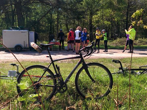 A black road bike parked on grass at a cycling rest stop with a group of cyclists in colorful jerseys gathered around a support vehicle and trailer in a wooded area during what appears to be an organized cycling event.