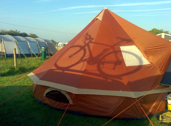An orange and cream colored camping tent pitched on grass at a campsite with other tents visible in the background. The tent appears to be a two-person model with guy ropes securing it to the ground. A bicycle wheel shadow is cast on the tent fabric, indicating the photo was taken from beside a parked bicycle.