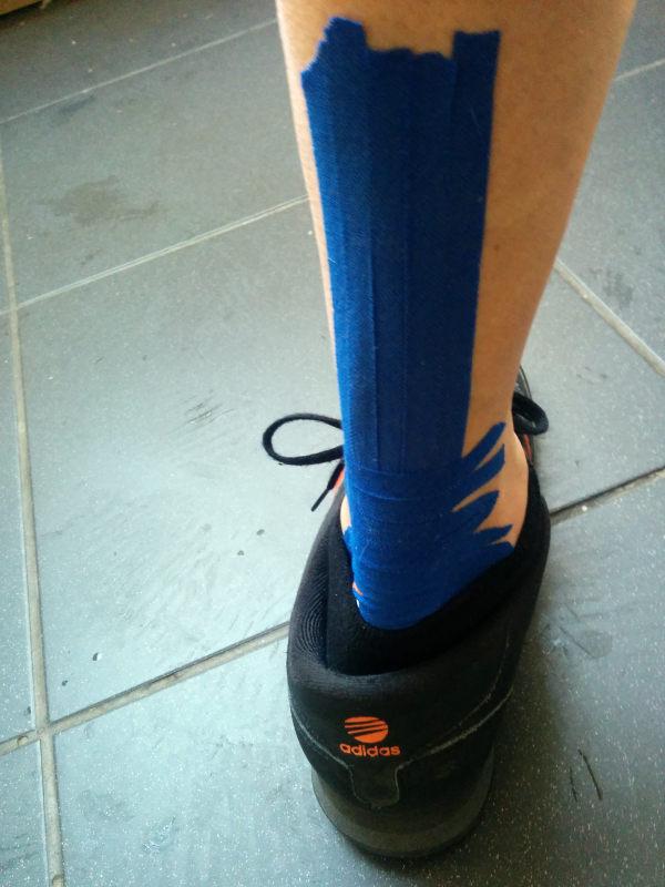 A close-up view of a cyclist’s lower leg and foot showing bright blue kinesiology tape applied from ankle to mid-calf, wearing a black Adidas training shoe on tiled flooring.