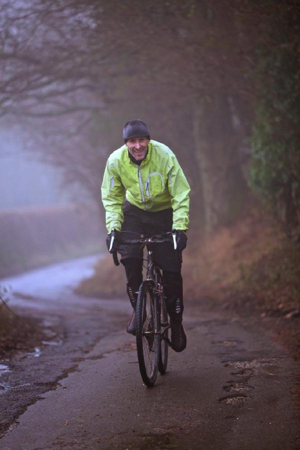 A cyclist in bright green high-visibility jacket and dark cap riding along a tree-lined country lane in misty conditions. The rider is on a dark colored bicycle and appears to be on a long-distance cycling event or audax ride, with atmospheric fog creating a moody backdrop through the overhanging trees.