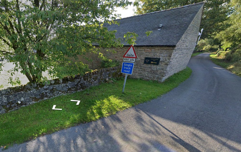 A narrow, winding country road curves past a stone building with a steeply sloped roof. In the foreground, a triangular road sign warns of a gate 650 yards ahead, with a blue sign below indicating a single track road. The building is made of rough stone and has a small sign on its wall. A low stone wall runs alongside the road, partially shaded by a leafy tree. The scene is quiet and rural, with greenery and trees surrounding the road and building.