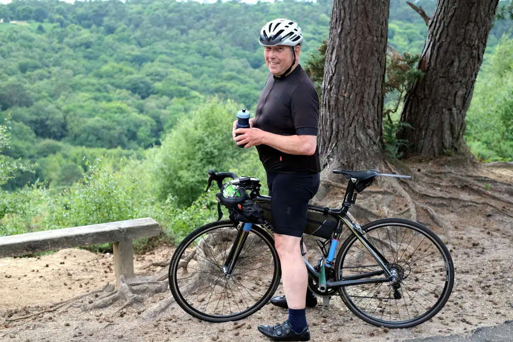 A cyclist stands next to a black road bike on a dirt path in a wooded area. The cyclist is wearing a white helmet, black cycling jersey, black cycling shorts, and dark socks with cycling shoes. He is holding a water bottle in both hands and facing slightly to the left. The background features dense green trees and rolling hills, with two large tree trunks rising behind him and a simple wooden bench to the left. The scene suggests a pause during a ride in a peaceful, natural setting.