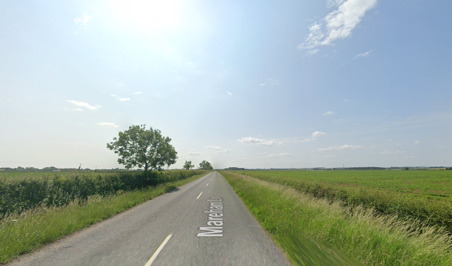 A straight, narrow country road called Mareham Lane stretches into the distance under a bright, mostly clear sky. The road is bordered by grassy verges and low hedges, with flat, open farmland on both sides. A few scattered trees line the roadside, and the landscape is expansive and rural, with no buildings in sight. The sun is high, casting a gentle light over the fields and the quiet scene.