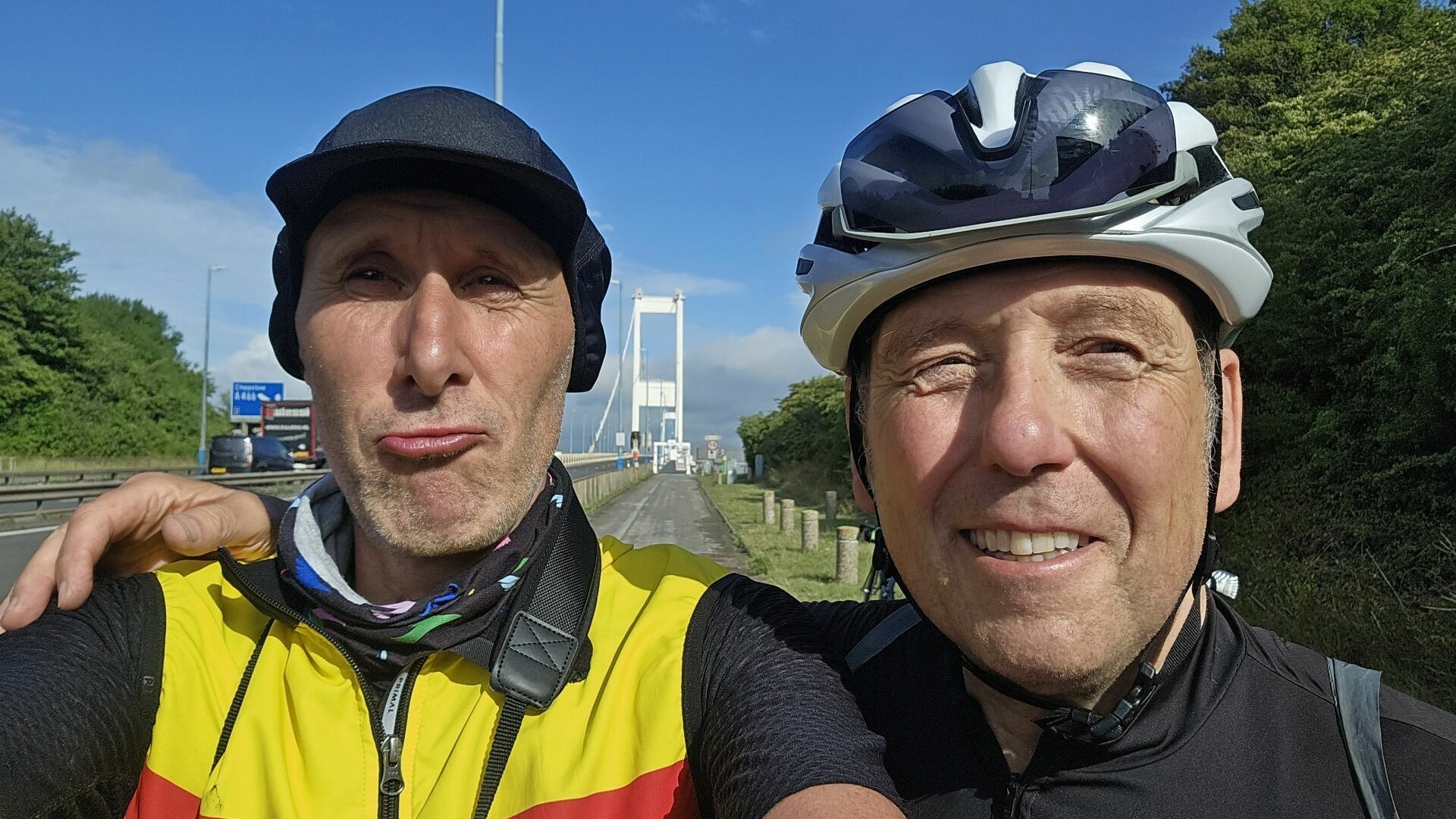 Two cyclists are taking a selfie on a sunny day. Both are wearing cycling helmets and gear; the cyclist on the left is in a yellow and red jacket, while the cyclist on the right is in black. They are standing on a path next to a road with cars visible on the left. Behind them, a large white suspension bridge stretches across the background, with green trees lining the right side of the image and a blue sky above. One cyclist has their arm around the other’s shoulder, suggesting camaraderie and enjoyment of their ride.