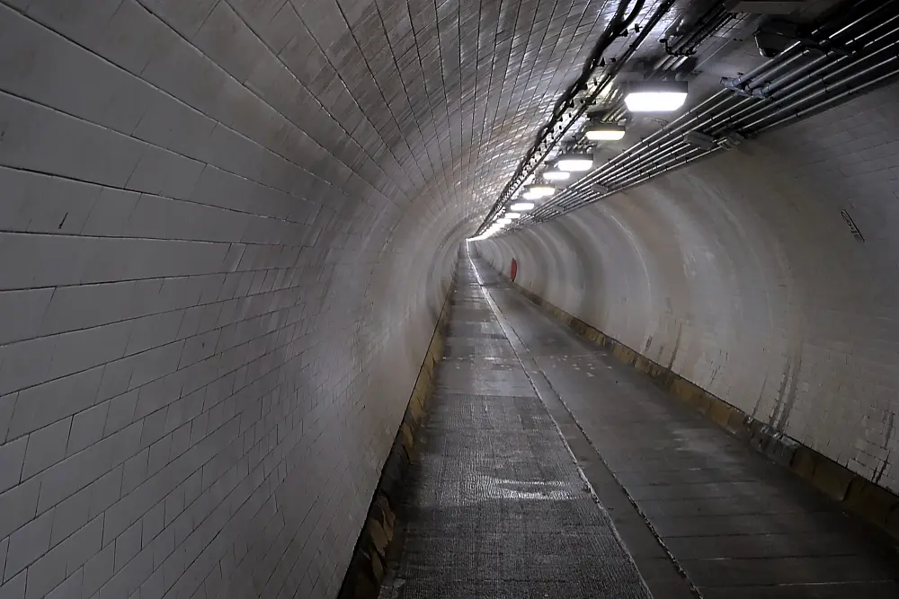 A long, curved underground pedestrian tunnel with white tiled walls and ceiling, illuminated by overhead lights, with utility pipes running along the right side near the ceiling. The floor is gently sloped, and the tunnel appears empty, stretching into the distance.