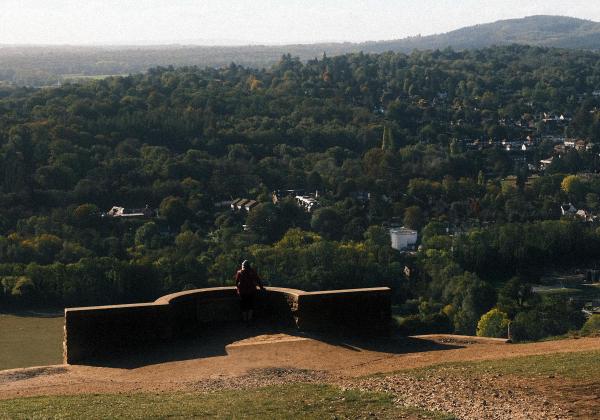 A person in red sits resting on a drystone wall at the top of a grassy hill, looking down over a town spread amongst trees, with hills fading into the distance in warm autumn light.